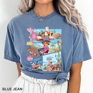 May include: Blue jean coloured t-shirt with a vibrant graphic from the animated film Zootopia. The design showcases characters such as Nick Wilde and Judy Hopps in a theme park scene. The text "UNDER SCUER MISSION" is also displayed.