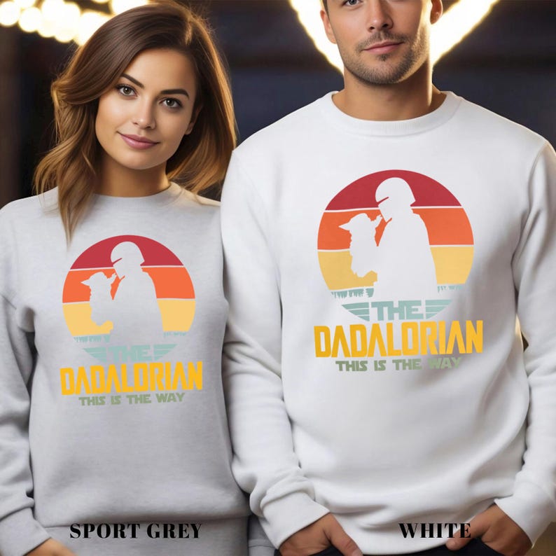 May include: A sport grey sweatshirt and a white sweatshirt, each featuring a graphic of a silhouette holding a small creature inside a sunset circle. Below the graphic, the text reads THE DADALORIAN and THIS IS THE WAY in yellow and green.