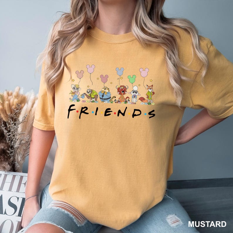 May include: Mustard-colored t-shirt featuring the word "FRIENDS" in black block letters. Above the word, there is a graphic of cartoon characters holding Mickey Mouse-shaped balloons. The shirt is a casual, unisex style.
