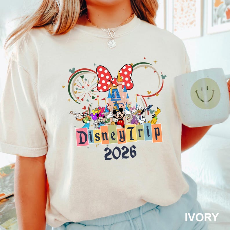 May include: Ivory-colored t-shirt featuring a colorful Disney graphic with Mickey Mouse, Minnie Mouse, and other characters. The design includes a castle, fireworks, and the words "Disney Trip 2026."