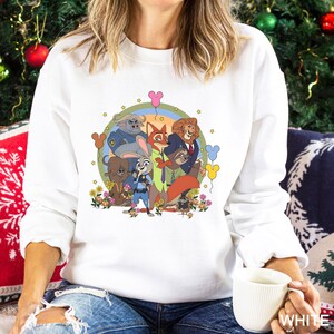 May include: White crewneck sweatshirt featuring a colorful graphic of animated animal characters. The design includes a rainbow, flowers, and Mickey Mouse-shaped balloons. The characters are surrounded by stars. The sweatshirt is a casual style.