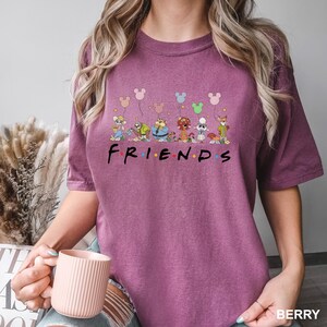 May include: Berry-colored t-shirt featuring the word "FRIENDS" in black letters. Above the word are cartoon characters and Mickey Mouse-shaped balloons. The shirt is a solid color and has short sleeves.
