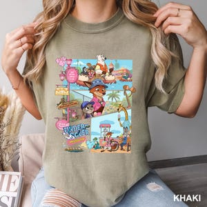 May include: Khaki t-shirt with a vibrant cartoon graphic. The graphic shows animal characters in a theme park, with the words "UNDER COVER MISSION". The shirt is made of a soft material.