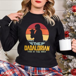 May include: A black long-sleeve sweatshirt featuring a graphic of a silhouette of a character holding a small creature against a sunset background. The text reads THE DADALORIAN THIS IS THE WAY in yellow and white lettering on the front of the garment.
