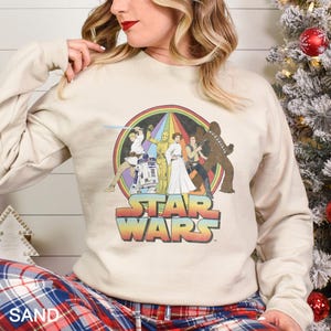 K&ouml;nnte beinhalten: Ein beiger Pullover mit einem Star Wars-Motiv im Vintage-Stil. Das Motiv zeigt Luke Skywalker, Prinzessin Leia, Han Solo, Chewbacca, R2-D2 und C-3PO in einem Regenbogenkreis mit dem Text "STAR WARS" darunter.