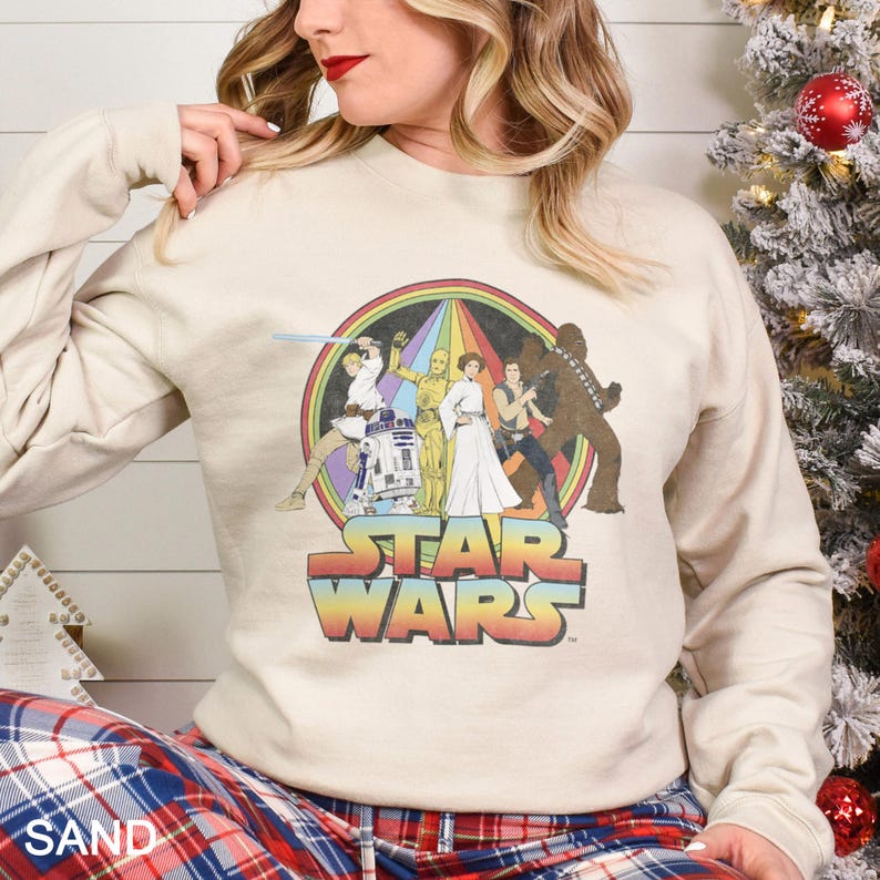 K&ouml;nnte beinhalten: Ein beiger Pullover mit einem Star Wars-Motiv im Vintage-Stil. Das Motiv zeigt Luke Skywalker, Prinzessin Leia, Han Solo, Chewbacca, R2-D2 und C-3PO in einem Regenbogenkreis mit dem Text "STAR WARS" darunter.
