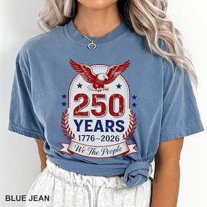 May include: A blue jean colored t-shirt with a graphic commemorating 250 years, from 1776 to 2026. The design features a red eagle, stars, and the text "We The People."