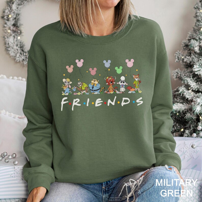 May include: An olive green sweatshirt featuring a cartoon design of various animal characters holding Mickey Mouse-shaped balloons above the word "FRIENDS" in white. The sweatshirt is a crew neck style and is made of a soft material.