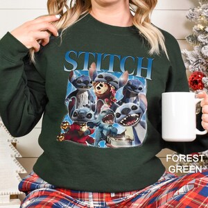 Op de afbeelding: Bostgroen sweatshirt met een afbeelding van Stitch uit Lilo & Stitch. Het woord "STITCH" staat in blauw boven de karakterillustraties. Een witte mok wordt in de hand van de persoon gehouden.