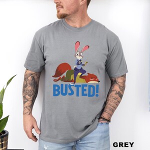 May include: Grey t-shirt featuring a cartoon image of a rabbit police officer standing on a fox, with the word "BUSTED!" in blue. The shirt is made of a soft material and is a unisex style.