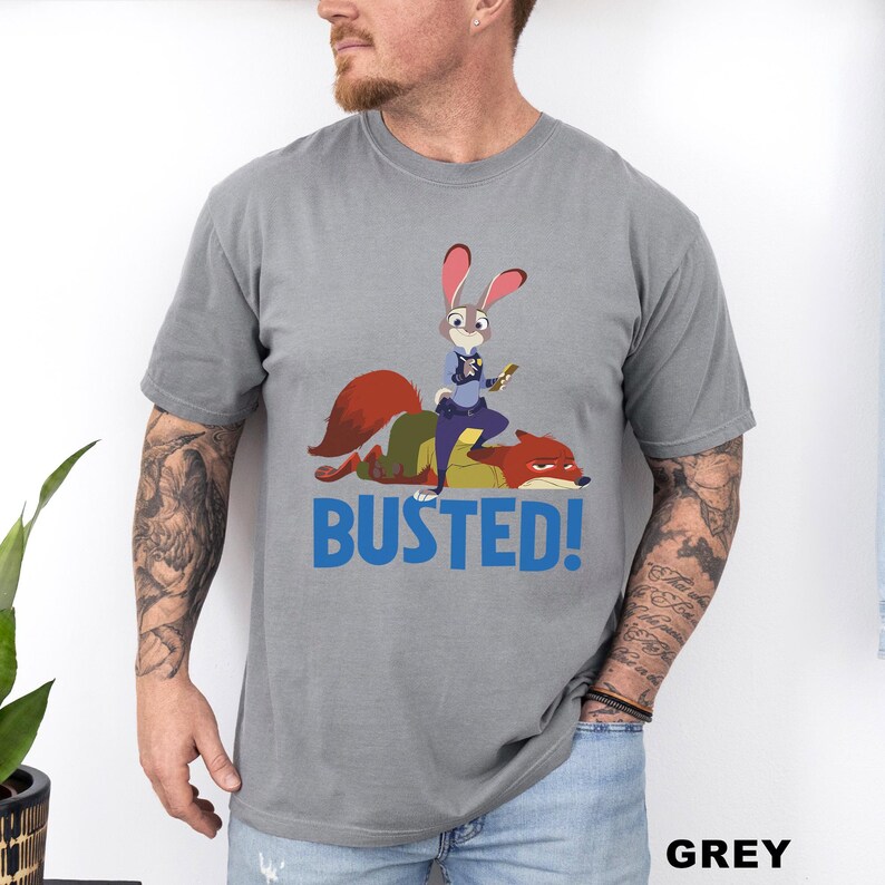 May include: Grey t-shirt featuring a cartoon image of a rabbit police officer standing on a fox, with the word "BUSTED!" in blue. The shirt is made of a soft material and is a unisex style.