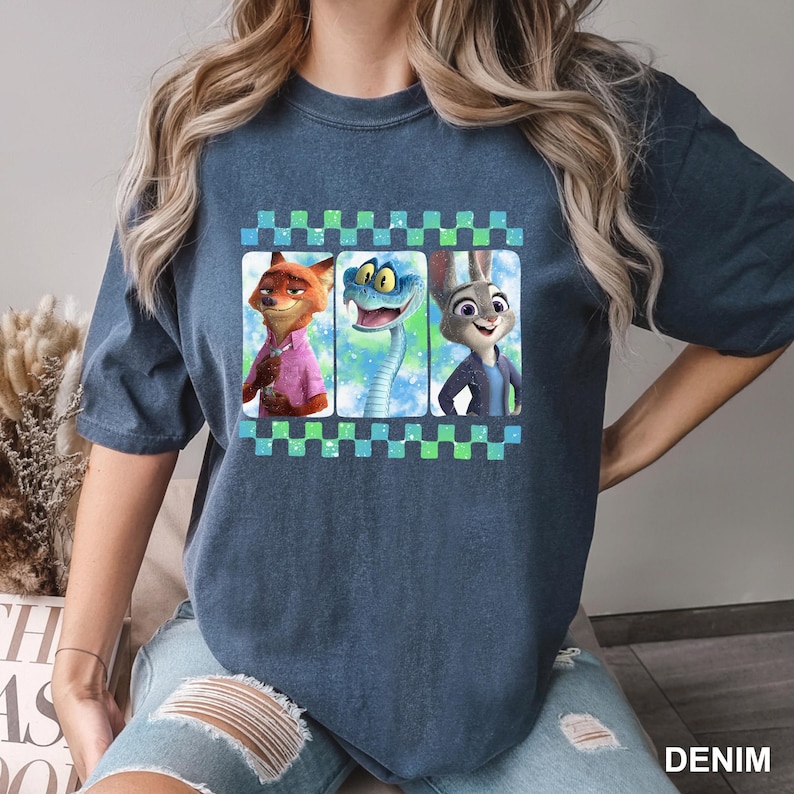 May include: A denim blue t-shirt featuring a graphic of three animated characters: a fox in a pink shirt, a snake, and a rabbit in a blue jacket. The graphic is framed with a checkered pattern.