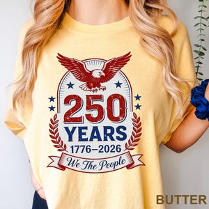 May include: Butter yellow t-shirt with a patriotic design. The graphic features a red eagle, blue stars, and the text "250 YEARS 1776-2026 We The People" in red and blue. The shirt is a comfortable, casual style.