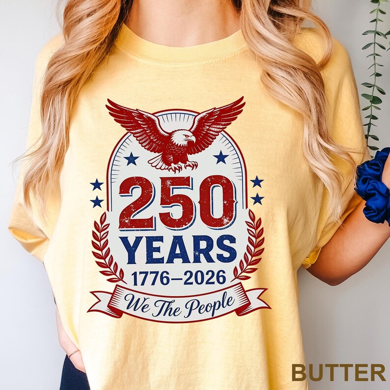 May include: Butter yellow t-shirt with a patriotic design. The graphic features a red eagle, blue stars, and the text "250 YEARS 1776-2026 We The People" in red and blue. The shirt is a comfortable, casual style.