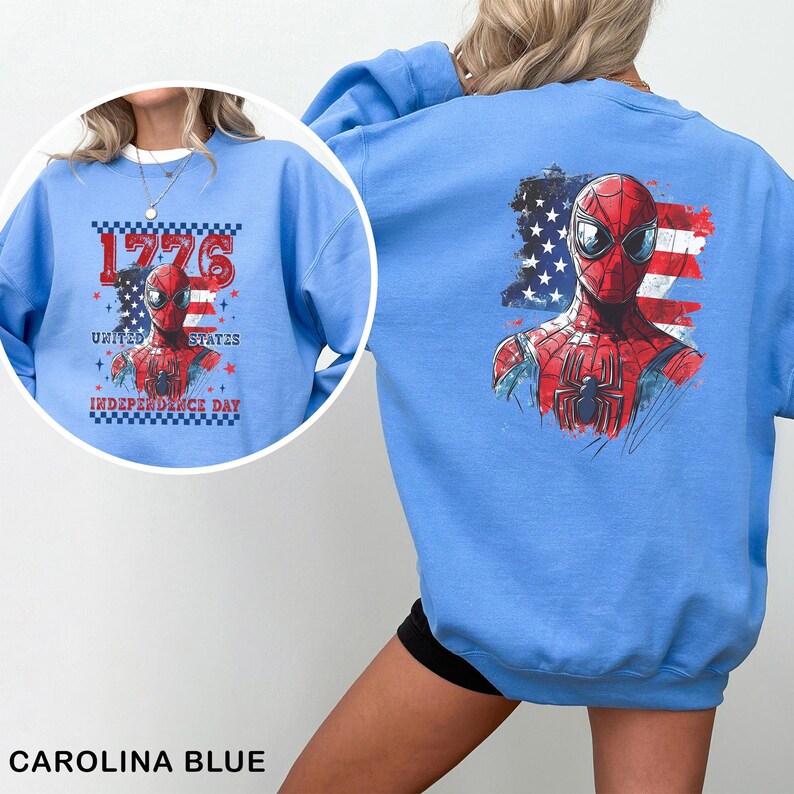May include: A light blue crewneck sweatshirt featuring a graphic of a red and blue superhero mask over a distressed American flag. The front design includes the text 1776 UNITED STATES INDEPENDENCE DAY with a checkered border and small stars.