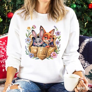 Puede incluir: Sudadera blanca con un gr&aacute;fico colorido de Judy Hopps y Nick Wilde de Zootopia, enmarcado por flores y placas de polic&iacute;a. La sudadera tiene mangas largas y cuello redondo. La palabra "WHITE" est&aacute; impresa en la parte inferior derecha.
