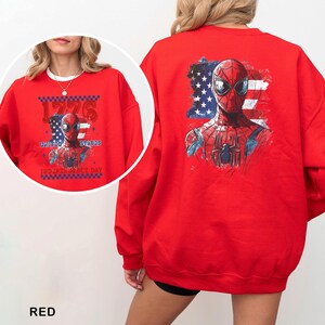 May include: A bright red crewneck sweatshirt featuring a graphic of the superhero Spider-Man layered over a distressed American flag. The design includes the text 1776, UNITED STATES, and INDEPENDENCE DAY in a bold, blocky font with a checkered border.