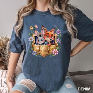 May include: Denim blue t-shirt with a vibrant illustration of Judy Hopps and Nick Wilde from Zootropolis, surrounded by flowers and police badges. The word "DENIM" is printed in white on the bottom right corner.
