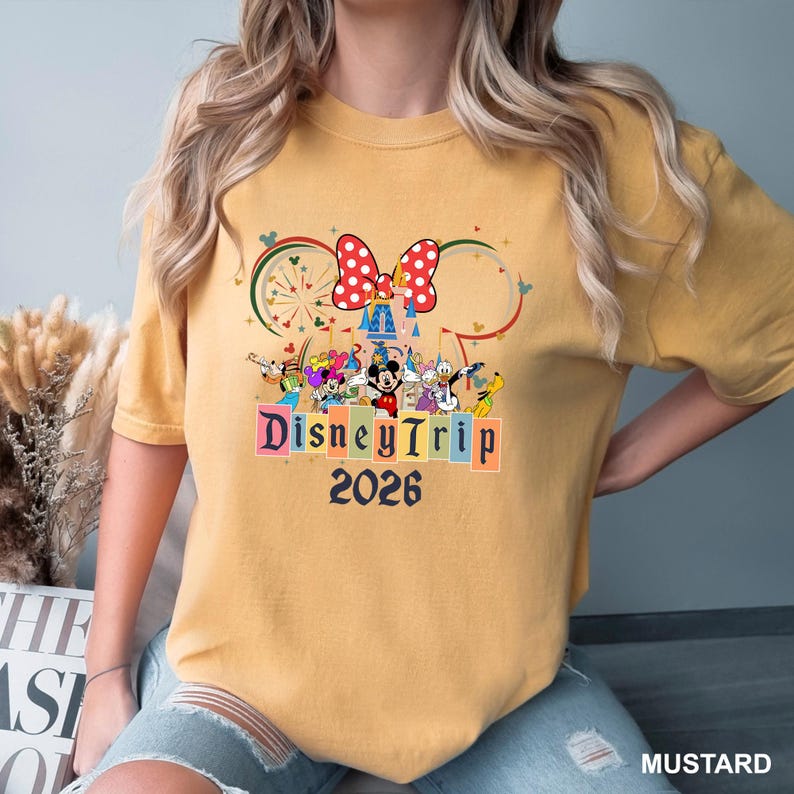 May include: Mustard-colored t-shirt featuring a Disney-themed graphic. The design includes Mickey Mouse, Minnie Mouse, and other characters, along with the words "DisneyTrip 2026". A red and white polka-dot bow is at the top.
