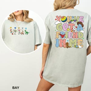 May include: Pale green t-shirt with a vibrant graphic of Toy Story characters on the front and the text "You've Got a Friend in Me" on the back. The design includes a rocket, cowboy hat, and other playful elements.