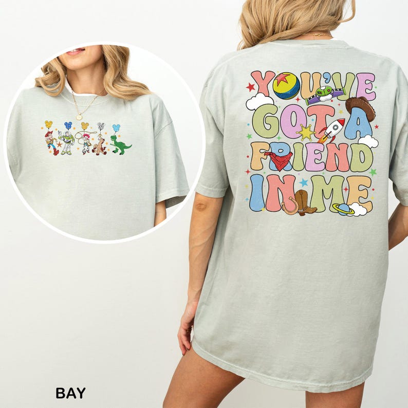 May include: Pale green t-shirt with a vibrant graphic of Toy Story characters on the front and the text "You've Got a Friend in Me" on the back. The design includes a rocket, cowboy hat, and other playful elements.