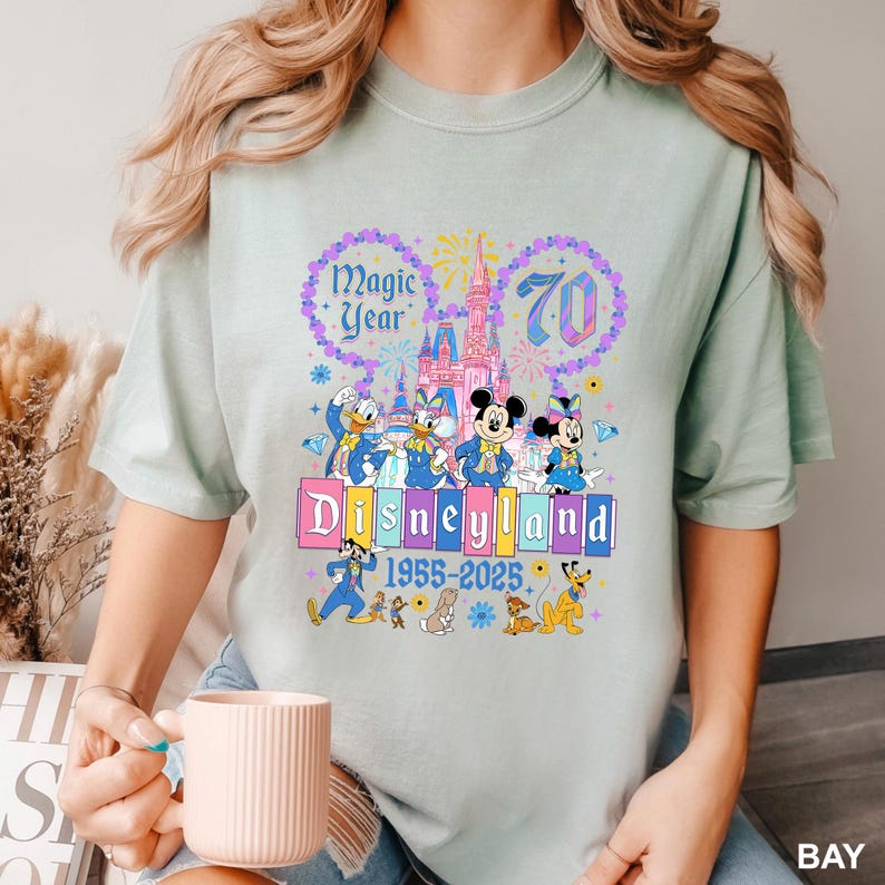 May include: A light green t-shirt featuring a colorful Disneyland graphic. The graphic includes Mickey and Minnie Mouse, Donald and Daisy Duck, and Goofy. The text reads "Magic Year 70 Disneyland 1955-2025".