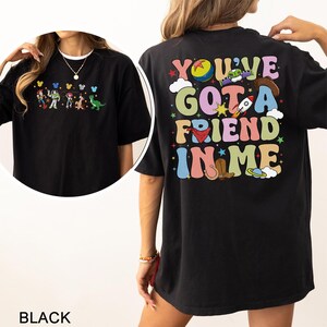 May include: Black t-shirt with a vibrant graphic of cartoon characters and the text "YOU'VE GOT A FRIEND IN ME." The front of the shirt shows a line of cartoon characters. The design includes a ball, a rocket, a cowboy hat, and a boot.