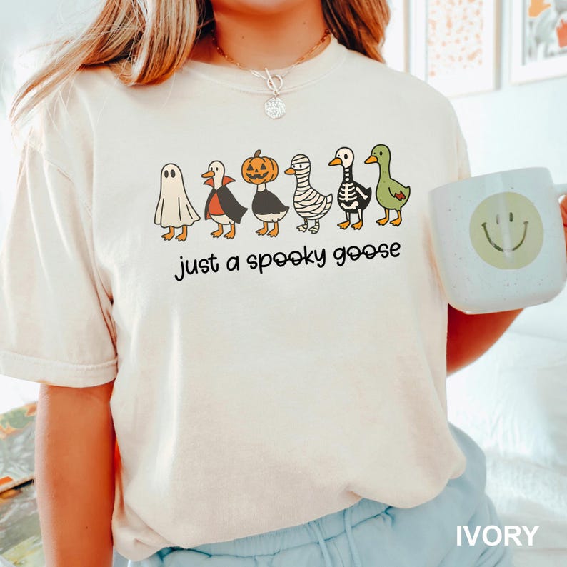 May include: Ivory t-shirt with a Halloween-themed design. It features cartoon geese in costumes: a ghost, vampire, pumpkin, mummy, skeleton, and green goose. The phrase "just a spooky goose" is printed below. A mug with a smiley face is on the right.