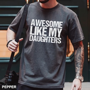 May include: Dark gray t-shirt with the text "AWESOME LIKE MY DAUGHTERS" in white. The shirt is a crew neck style and has short sleeves. The word "PEPPER" is at the bottom left of the image.