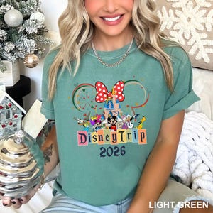 May include: Light green t-shirt featuring a colorful Disney Trip design. The graphic includes a castle, Minnie Mouse bow, and various Disney characters. The text "Disney Trip 2026" is displayed in colorful block letters.