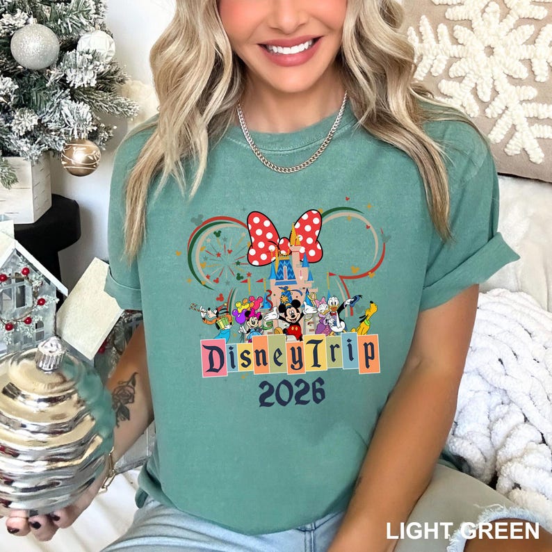 May include: Light green t-shirt featuring a colorful Disney Trip design. The graphic includes a castle, Minnie Mouse bow, and various Disney characters. The text "Disney Trip 2026" is displayed in colorful block letters.