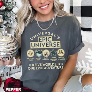 May include: Dark gray t-shirt with the text "UNIVERSAL'S EPIC UNIVERSE" and icons representing different theme park areas. The shirt also says "FIVE WORLDS. ONE EPIC ADVENTURE."