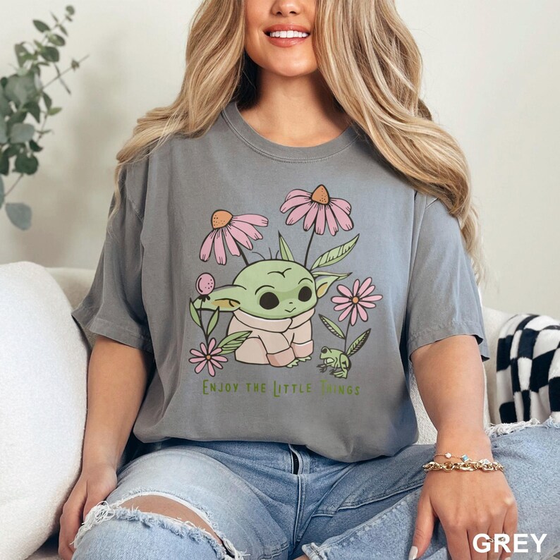 May include: A grey t-shirt featuring a cartoon illustration of a green alien character with large ears and a pink shirt, surrounded by pink flowers and a green frog. The text "Enjoy the little things" is printed below the image.