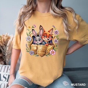 May include: Mustard-coloured t-shirt with a cartoon image of Judy Hopps and Nick Wilde from Zootopia, framed by flowers and a police badge. The shirt is a comfortable, casual style.
