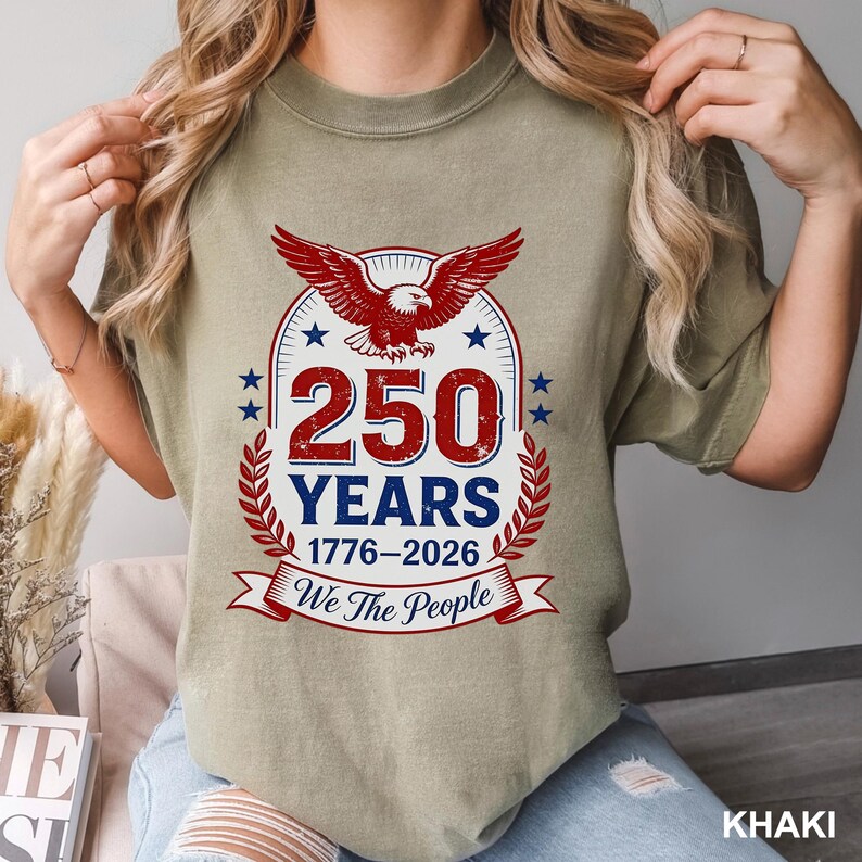 May include: Khaki-colored t-shirt featuring a patriotic design. The graphic includes a red eagle, blue stars, and the text "250 YEARS 1776-2026 We The People" in red and white. The shirt is a casual style.
