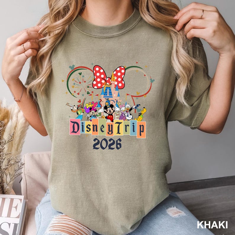 May include: Khaki-colored t-shirt featuring a colorful Disney Trip 2026 graphic. The design includes Mickey Mouse, Minnie Mouse, and other Disney characters. A large red bow with white polka dots is at the top of the design.