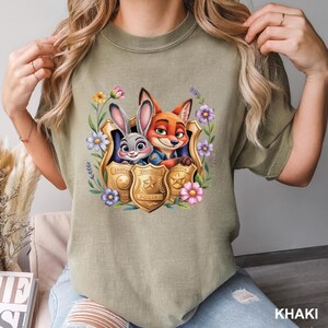 May include: Khaki t-shirt with a colourful graphic of Judy Hopps and Nick Wilde from Zootopia, surrounded by flowers and police badges. The text "Zootopia Police" is on the badges. The shirt is a comfortable, casual style.