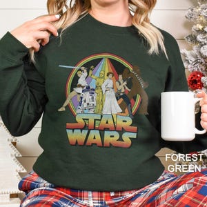 K&ouml;nnte beinhalten: Ein dunkelgr&uuml;nes Sweatshirt mit einem Vintage-Star-Wars-Motiv, das Luke Skywalker, Prinzessin Leia, Han Solo, Chewbacca, R2-D2 und C-3PO zeigt. Das Motiv ist von einem Regenbogen umgeben.