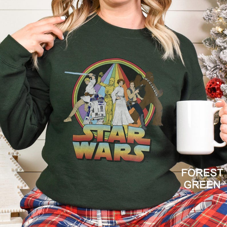 K&ouml;nnte beinhalten: Ein dunkelgr&uuml;nes Sweatshirt mit einem Vintage-Star-Wars-Motiv, das Luke Skywalker, Prinzessin Leia, Han Solo, Chewbacca, R2-D2 und C-3PO zeigt. Das Motiv ist von einem Regenbogen umgeben.