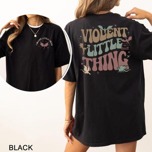 May include: Black t-shirt with the text "VIOLENT LITTLE THING" in a retro font on the back and front. The design includes dragon illustrations and stars. The shirt is a casual, oversized fit.