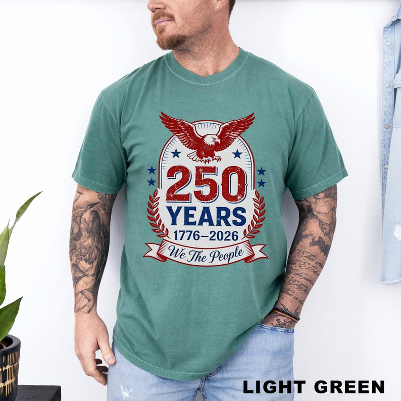 May include: Light green t-shirt featuring a patriotic design. The graphic includes a red eagle, the number "250" in red, and the text "YEARS 1776-2026 We The People" in a circular emblem with stars and laurel leaves.