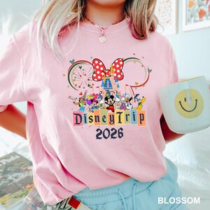 May include: Pink t-shirt with a colorful Disney Trip 2026 graphic featuring Mickey Mouse, Minnie Mouse, and other Disney characters. The design includes a large red bow with white polka dots and a castle.