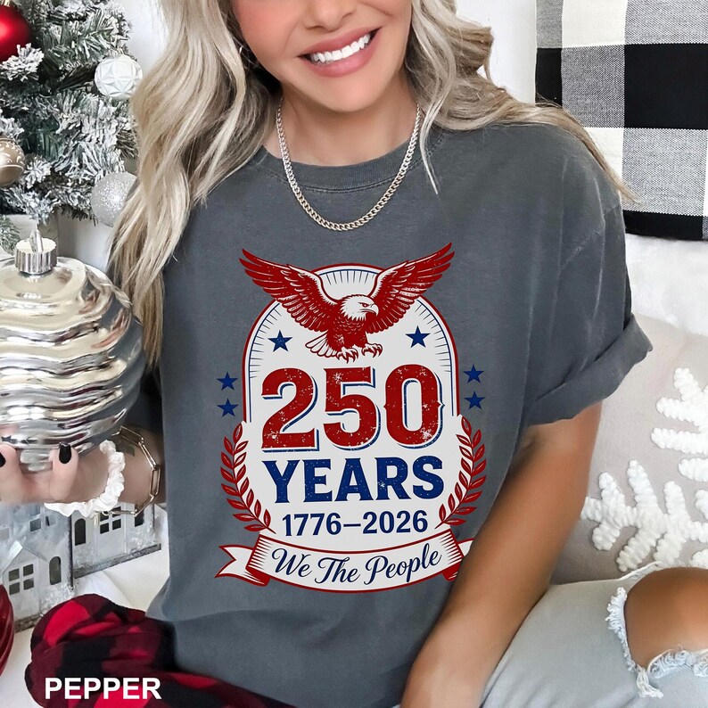 May include: A gray t-shirt with a patriotic design. The graphic features a red eagle, the number "250" in red, and the words "YEARS 1776-2026 We The People" in white. The design is framed by a wreath and stars.