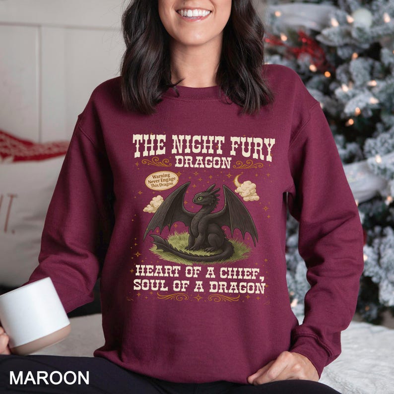 Puede incluir: Sudadera granate con un dise&ntilde;o de un drag&oacute;n negro con ojos verdes y el texto "THE NIGHT FURY DRAGON". El dise&ntilde;o incluye el texto "HEART OF A CHIEF, SOUL OF A DRAGON" y una advertencia.