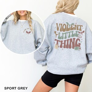 May include: Sport grey sweatshirt with the text "VIOLENT LITTLE THING" on the back in brown, green, and red. The front of the sweatshirt has a smaller design with the text "VIOLENT LIT LIL" and a dragon graphic.