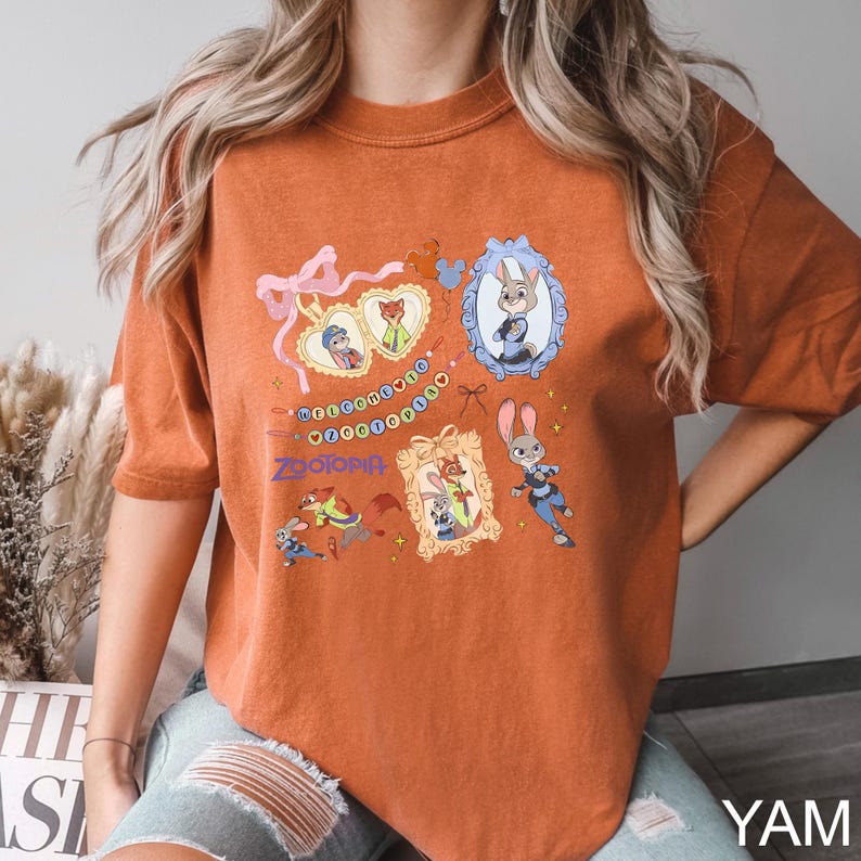 May include: Burnt orange t-shirt featuring multiple framed illustrations of characters from the animated film "Zootopia." The design includes the word "Zootopia" and various character images, with a decorative string of beads.