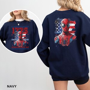 May include: A navy blue crewneck sweatshirt featuring a graphic of a red and blue superhero mask over a distressed American flag. The front design includes the text 1776, UNITED STATES, and INDEPENDENCE DAY in red and blue lettering.