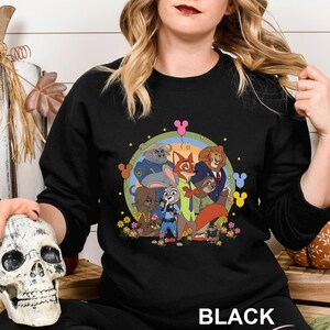 May include: Black crewneck sweatshirt featuring a colorful graphic of characters from the animated movie Zootopia. The design includes Judy Hopps, Nick Wilde, and other characters, surrounded by flowers and stars. The word "BLACK" is printed at the bottom.
