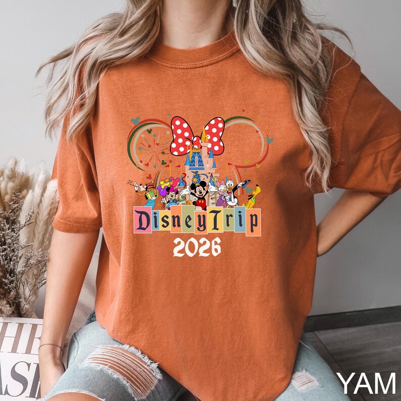 May include: Burnt orange t-shirt featuring a colorful graphic with Disney characters, a castle, and a polka-dot bow. The text "DisneyTrip 2026" is displayed below the graphic. The shirt has short sleeves and a relaxed fit.
