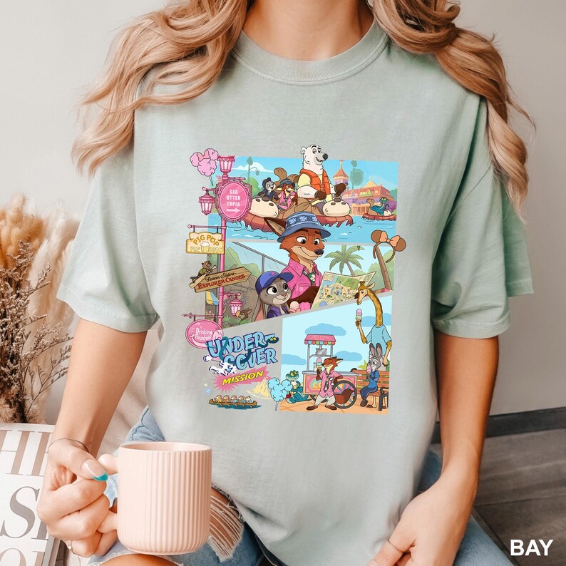 May include: A pale green t-shirt with a vibrant cartoon illustration of animal characters in a theme park setting. The design includes the words "Undercover Mission" and other signs. The shirt is plain and has short sleeves.